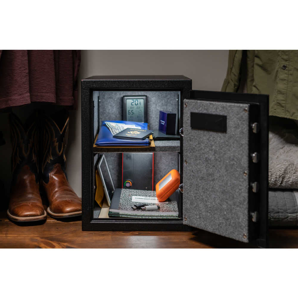 Hornady 95407 Fireproof Keypad Safe with its door open, showing a gray carpeted interior with shelves holding documents, a wallet, digital thermometer, and other valuables. The safe is displayed on a wooden floor in a home setting.