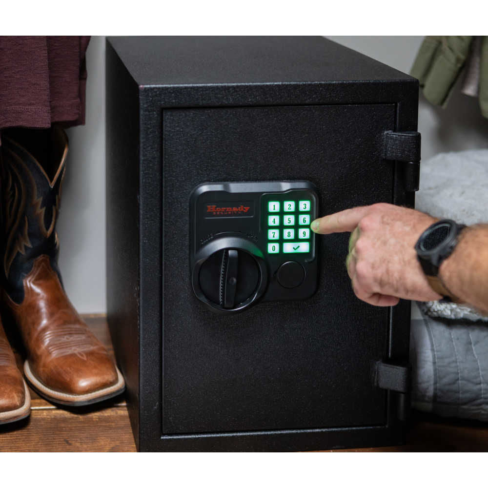 Hornady 95407 Fireproof Keypad Safe front view A black electronic safe with a keypad and thumb turn lock, shown with a person's hand pressing numbers on the keypad. The safe is positioned next to a person wearing brown cowboy boots, with a gray wall and bedding in the background.